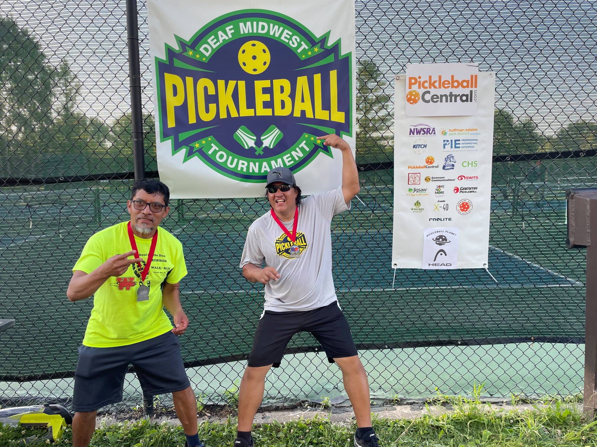 Carlos Lopez and Wilson Velazquez standing in front of Deaf Midwest Pickleball logo.